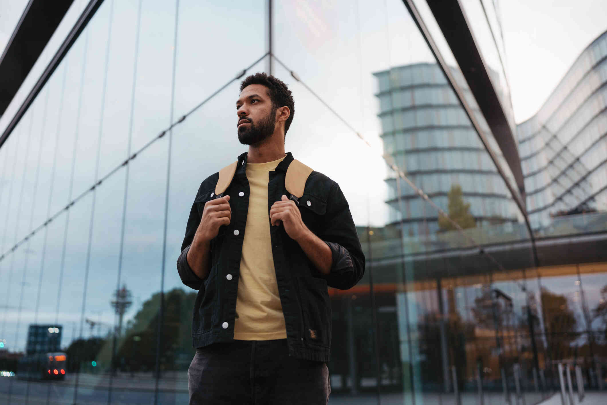 A man with abrown backpack stands outside of a large glass building in the city and gazes off with a serious expression.